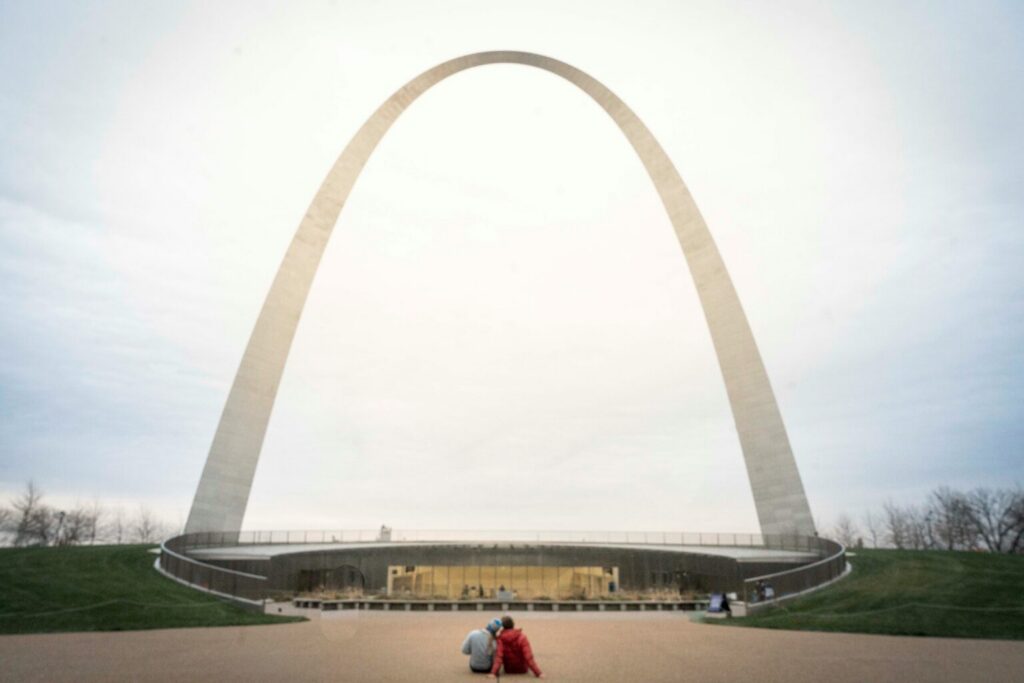 Two people seated looking at an arch building in the daytime.