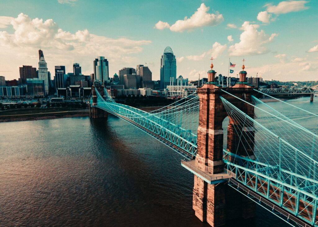 Elevated aerial view of the historic blue Roebling Suspension Bridge spanning the wide brown Ohio River, leading directly to the dense Cincinnati, Ohio skyline filled with modern skyscrapers under a bright blue sky. This view captures the heart of the bustling, established business district, a thriving economic environment influenced by factors like the Ohio corporate tax rate.