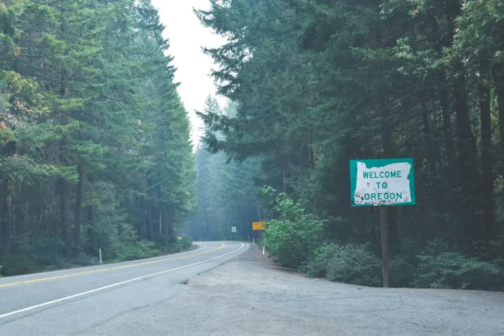 An aerial-style view of a road leading through a dense, evergreen forest with a "Welcome to Oregon" sign on the shoulder. This gateway signifies the point where businesses must begin navigating the Oregon corporate tax rate and its unique state-level compliance requirements.