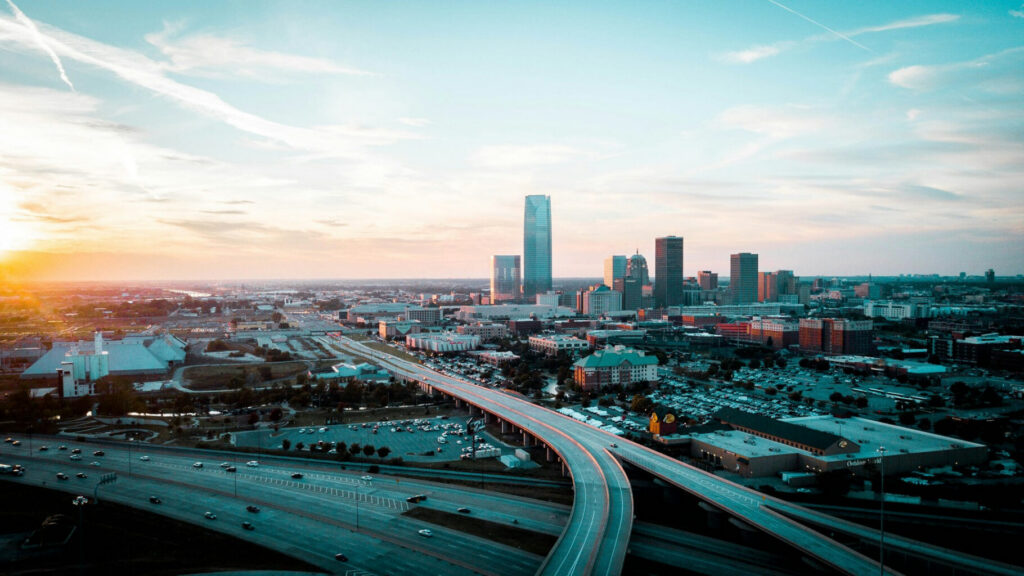 An expansive aerial sunset view of the Oklahoma City skyline and major highway interchanges. The modern skyscrapers of the city center represent a growing business environment, a key factor for companies monitoring the Oklahoma corporate tax rate.