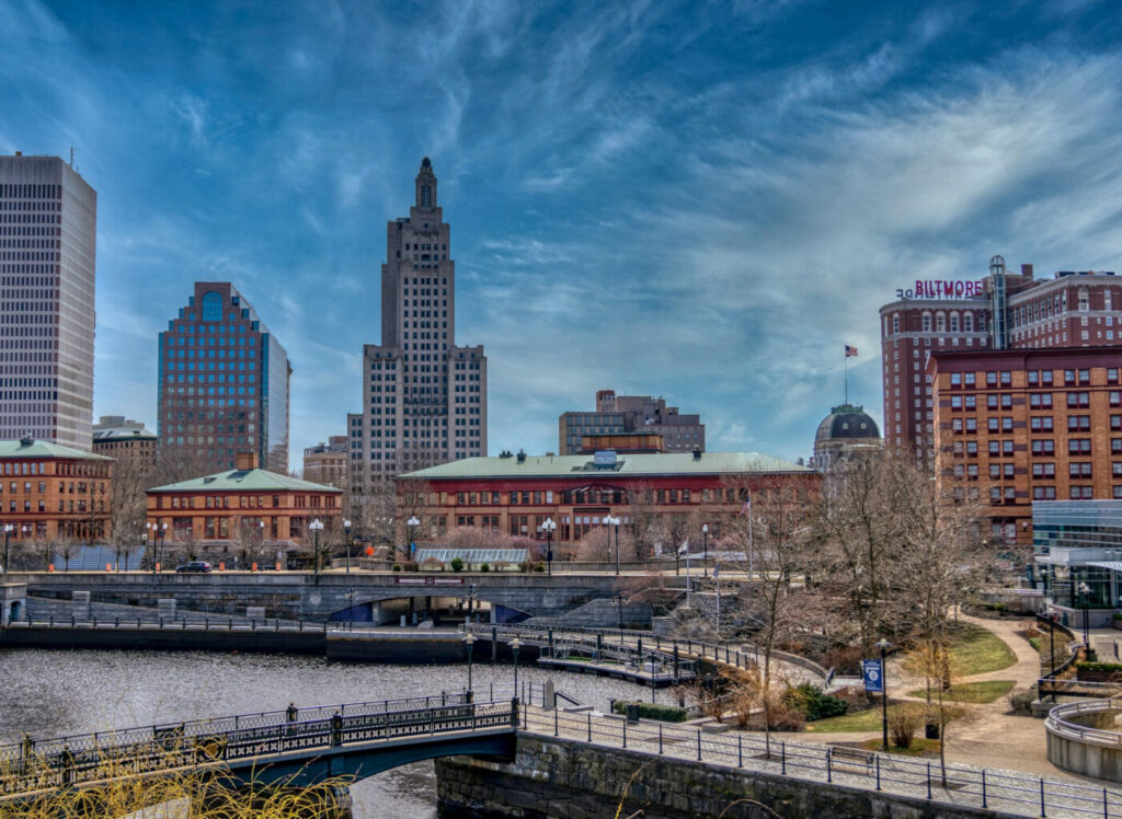 A scenic view of the Providence, Rhode Island skyline under a vibrant blue sky with wispy clouds, featuring the historic Industrial Trust Tower and the Biltmore Hotel, overlooking a river with pedestrian bridges and landscaped walkways.