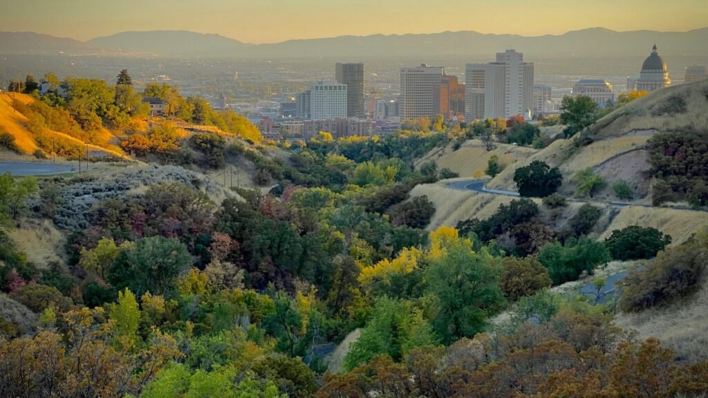View of the Salt Lake City skyline from tree-covered foothills at golden hour, with the Utah State Capitol visible on the right and mountain ranges in the distance