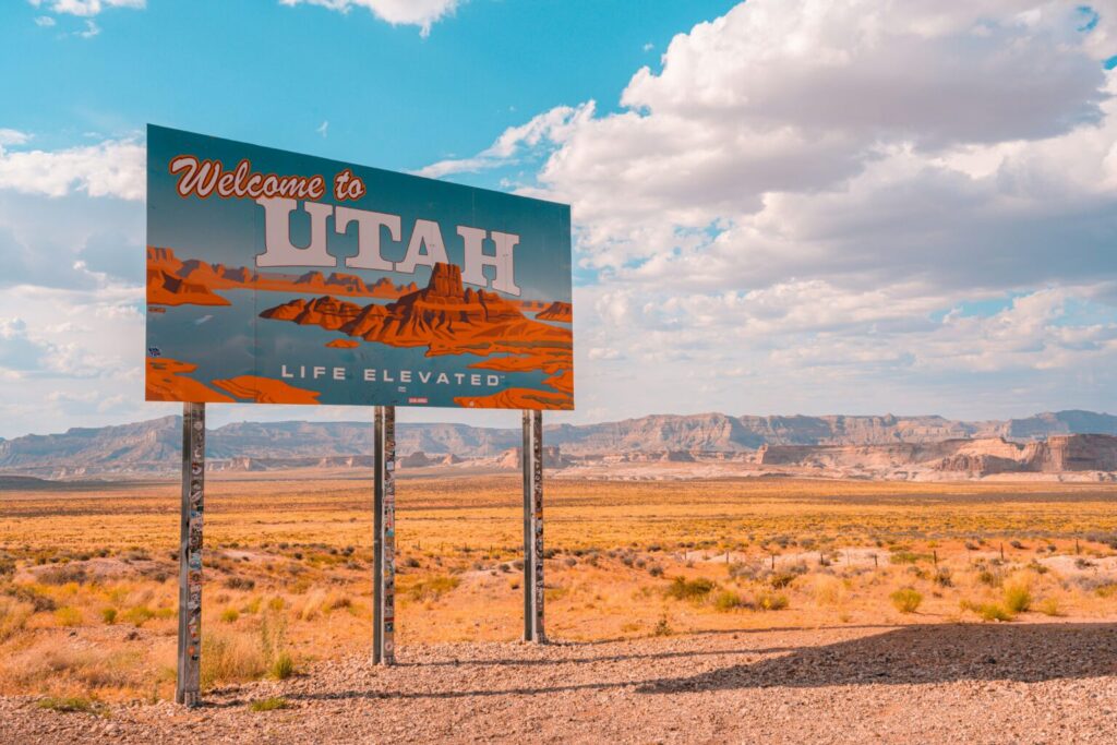 Welcome to Utah road sign reading Life Elevated, set against an open desert landscape with red rock mesas and a bright blue sky