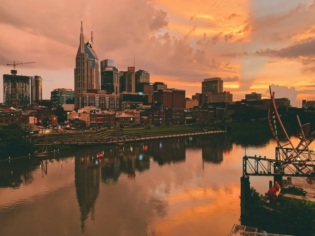 Aerial view of the Nashville city skyline at dusk, reflected in the Cumberland River with a vivid orange and pink sky overhead