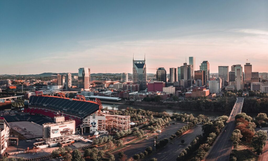 Aerial daytime view of the Nashville skyline with a large stadium in the foreground, the Cumberland River, and rolling hills in the background under a pale blue sky